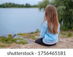 Backview of small girl sitting alone by the river. Peaceful lifestyle shot of enjoying beauty of nature. Relaxing outdoors on vacation.