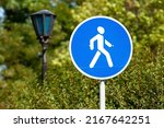 pedestrian zone road sign close-up on a background of park green trees. round blue road sign with white human icon. pedestrian zone in the park road sign next to a vintage street lamp.