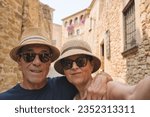 Small photo of Elderly couple taking a selfie with smartphone outdoors - Two retired tourists enjoying summer vacations together in the village of Pals, Girona,Catalonia - Lifestyle concept with mature woman and man