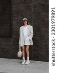 Small photo of beautiful blonde girl dressed in milky oversized jacket, short gray shorts, t-shirt, beige cap, massive boots, bag in hand, accessories, sunglasses, stylish fashion outfit, full length lifestyle model