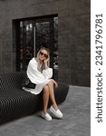 Small photo of beautiful blonde girl dressed in white light dress, loafers, black shoulder bag, accessories, sunglasses, stylish fashion outfit, full length lifestyle model sitting