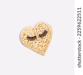 Small photo of English crumpet, heart shape with fake eyelashes, creative love inspired layout, white background.