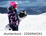 Little girl snowboarder with equipment helmet and goggles outwear holding snowboard resting on top of ski slope in sunlight. 6-7 years girl at ski resort in sunny winter day. Free space for you text