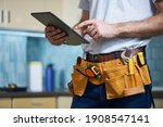Cropped shot of young repairman wearing a tool belt with various tools using digital tablet while standing indoors