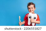 Small photo of A smiling boy with healthy teeth holds a plush tooth and a toothbrush on a blue isolated background. Oral hygiene. Pediatric dentistry. Prevention of caries. A place for your text.