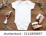Mockup of white baby bodysuit on dark wood background with greenery, booties and toy. Blank baby clothes template in farmhouse style, flat lay.