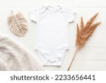 Mockup of white baby bodysuit on wood background with dried flowers, knitted plaid and macrame decor, flat lay. Blank baby clothes template in neutral color, boho style.