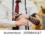 Small photo of A close-up of a Jewish bar mitzvah boy, wearing a white shirt and a red tie. wearing a tefillin for the first time in a synagogue