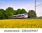 Small photo of Modern fast train passing a yellow blooming rapeseed field on the main railway track between Dortmund, Bochum and Essen in Ruhrbasin Germany. Colorful landscape on a sunny springtime day.