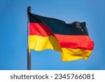 German flag in black-red-gold color on a sunny day in Germany. National yymbol of the “Bundesrepublik Deutschland“. Flag pole and wavy fabric blown in the wind on a blue sky day. Intense sunlit colors