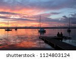 Amazing summer evening landscape with group of drifting yachts on a lake, two people on a wooden pier. Mendota during spectacular sunset. Bright sky reflects in the lake water.