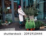 Young woman wearing protective medical mask in faux fur coat and hat picks small Christmas tree in wicker pot in city flower shop. Christmas market, preparation for holiday