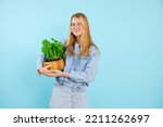 Nice teenage girl hold in hands green houseplant in wooden pot on empty blue background, free copy space. Photo of charming young lady growing indoor plant. Home flower grower, plant care.