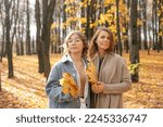 Small photo of Portrait of beauteous family of middle-aged woman mother and young teenage girl daughter standing closely near trees in park forest in sunny autumn, holding yellow maple leaves, enjoying sunlight.