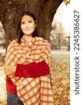 Small photo of Portrait of young beauteous woman with dark hair wearing vinous sweater, beige orange checkered warm scarf, jeans, standing near tree with crossed hands among yellow fallen leaves in forest park.