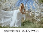 Small photo of Adorable young woman in wedding gown standing close to cherry blossom, leaning over branch of tree and smiling. High heels shoes of lady hang on ramification of sakura, bride going to take footwear.