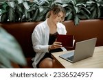 Young female freelancer in white shirt and black dress sitting on brown leather sofa at cafe table in front of laptop and holding diary. Woman with ponytail opening sketchpad while busy with job.