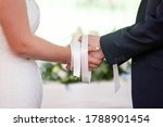 Wedding ceremony moment, bride and groom's hands tied with white ribbons, white wedding dress, dark suit
