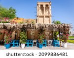 Traditional Arabic style building with windcatcher tower and palm leaves thatched roof; blue benches and pink flowers around, Old Dubai, UAE.