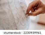 Morning preparation of the bride. Close-up of a bridesmaid, fastening a lot of buttons on the bride's wedding dress. The bride in a white wedding dress with lace standing in the room. Selective focus