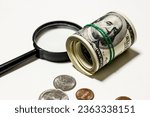 Hundred dollar bills are rolled up with rubber band with coins and magnifying glass on white background