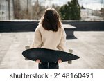Young female standing back with sport skateboard on windy rooftop. Woman with dark hair in outwear clothes enjoying street activity and beautiful view from high building.