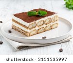 Tiramisu cake decorated with cocoa powder and fresh green mint leaf on white ceramic plate. Close up food. Traditional italian dessert. Coffee beans, textile napkin, white wooden table background.