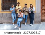 Small photo of Five young, multiracial friends in urban attire, leaning on a city wall, absorbed in their mobile phones, oblivious to their surroundings.