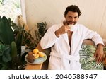 Handsome man drinking wellness tea while relaxing at exotic spa resort, wearing a white bathrobe and sitting on armchair
