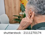 Small photo of Hearing solutions for elderly deafness people. Older gray-haired man tunes his hearing aid behind the ear by pressing his finger on setting button while visit to hearing clinic