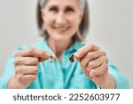 Small photo of Mature woman with hearing problems chooses between behind-the-ear hearing aid and in-the-ear hearing aid by holding them in front of her. Hearing aids and solutions