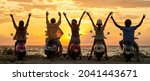 Back view of cheerful young friends, motorcycle riders raised their hands while standing with their scooters on the beach and watching sunset together. Friendship, nature concept. Rear view