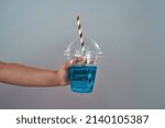 Small photo of Cropped image of little girl hand holding plastic cup with tubule and soda drink. Blue fresh sweet drink. Childhood concept. Isolated on grey background. Studio shoot. Copy space