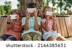 Small photo of Obscure faces of children using and watching smartphones on wooden bench in park outdoors. Boys and girl of generation alpha. Gadget addiction. Childhood lifestyle. Sunny summer day
