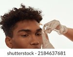 Small photo of Doctor hands wearing latex gloves making injection of botox with syringe on face of black guy. Obscure face of y young curly man. Face lifting and mesotherapy. Isolated on white background in studio