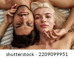 Partial view of serious young beautiful multiethnic couple lying and looking at camera on bed at home. Caucasian girl and middle eastern man touching each other. Domestic lifestyle. Closeness