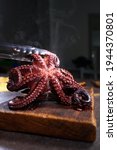 Small photo of Boiled whole octopus on a wooden cutting board. The octopus is held with tongs. Cooking seafood dishes. Dark background. Protein food. Free place. Vertical photo.