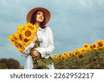 Lovely woman hold yellow bouquet bunch blooming sunflower field outdoors sunrise warm nature background. Smiling lady dressed white shirt wear hat posing standing outside, agriculture concept
