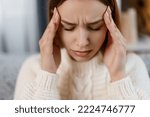 Small photo of Close up portrait of displeased caucasian woman suffering from migraine while staying alone at cozy apartment. Unhappy young blonde sitting on couch and massaging temples.