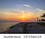 Small photo of A colorful sky during the golden hour with clouds around the sun. A walking path with a stone chair appear alongside the sea (corniche), in Dareen, Tarout Island, Saudi Arabia.