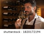 Authentic shot of successful male sommelier is tasting a flavor and checking white wine quality poured in transparent glass in a wine cellar.
