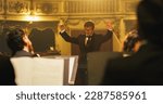Small photo of Cinematic Close Up of Conductor Directing Symphony Orchestra with Performers Playing on Stage During Music Concert. Professional Conductor Leading Musicians in Classic Theater