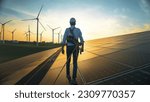 Small photo of Professional Male Green Energy Engineer Walking On Industrial Solar Panel, Wearing Safety Belt And Hard Hat. Man Inspecting Sustainable Energy Farm With Wind Turbines On Background.