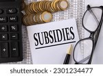 Small photo of SUBSIDIES - word on a white piece of paper on the background of a calculator, pennies and glasses