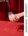 Small photo of Woman hand holding glass and pouring wine of bottle. Woman tasting white wine on red background. Minimal trendy style composition with person and champagne. Pouring wine and female hand with glass