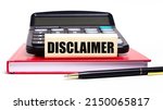 Small photo of A burgundy notebook on a white background. On it is a black calculator, a pen and a wooden block with an DISCLAIMER text. Business concept