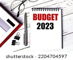 Small photo of Notepad with text BUDGET 2023, glasses, paper clips, white calculator and pen on desktop, top view.