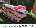 Small photo of Old furniture disjointed on a pile with a big pink teddy bear lying prone on it. The pile consists mostly of bed construction, grate and mattress covered with white sheets. The pile is on a lawn.