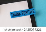 Small photo of Close-up of a blue sticky note with THUNK POSITIVE on it, attached to a black and white notebook. A black pen is placed on top. Set against a well-lit blue background, isolated for emphasis.