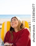 Small photo of Young woman smiling while sitting in front of the beach during a sunny day. Girl enjoying life in a terrace very happy. Woman with a phone laughing after recieve a message, chuffed.